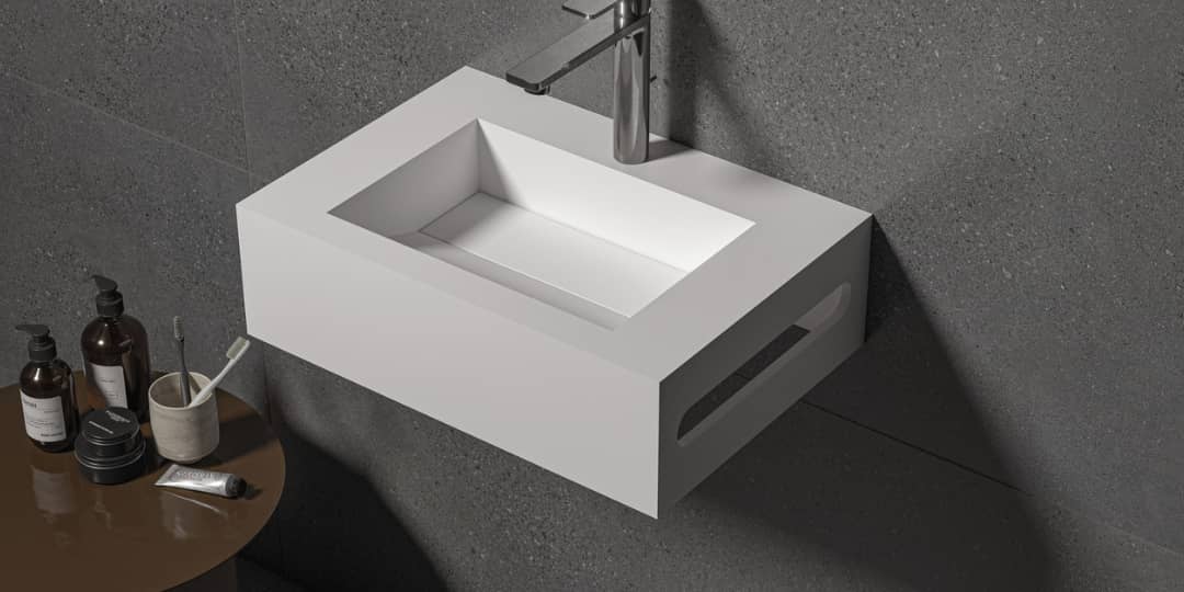 Lavabo suspendido modelo Ariel en color blanco, fabricado en material Solid Surface, instalado en baño moderno con pared gris oscura.