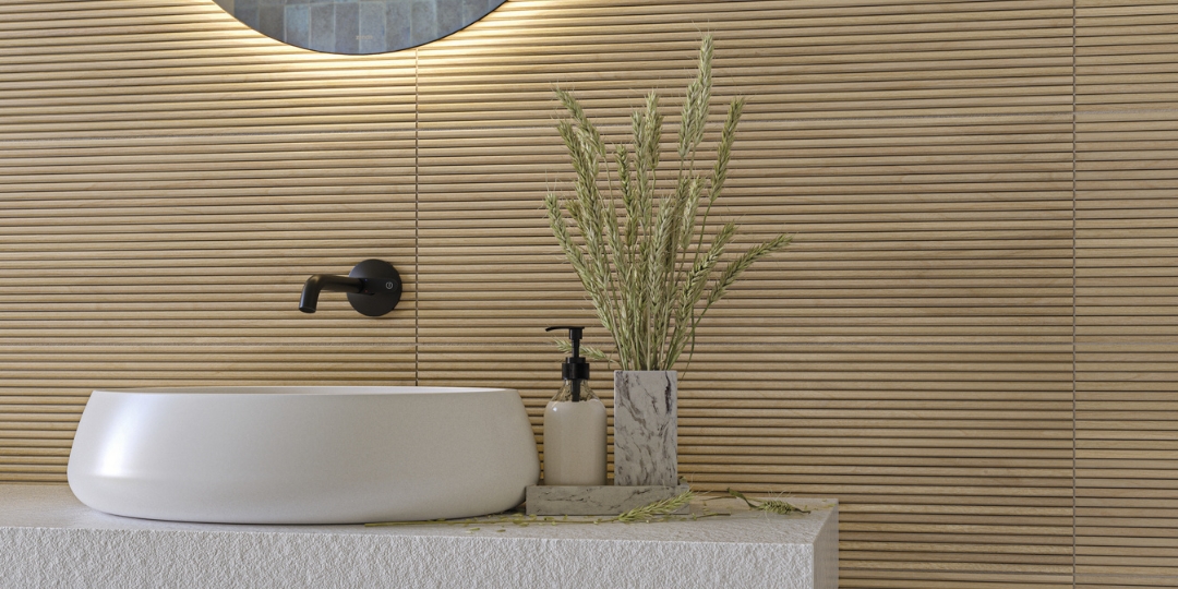 Lavabo redondo blanco sobre encimera texturizada, con grifería negra de pared y decoración natural en tonos cálidos.

