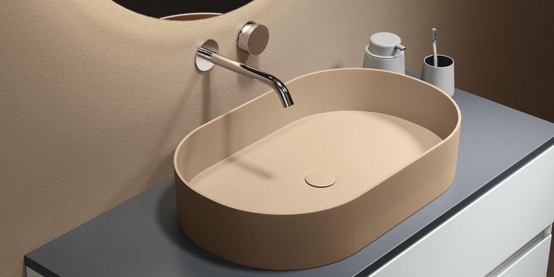 Lavabo de diseño ovalado en color beige con grifería cromada empotrada en pared, sobre encimera gris en baño contemporáneo.