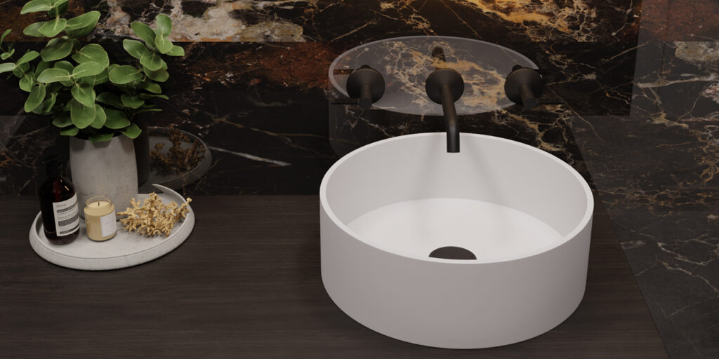 Lavabo redondo blanco modelo Diana instalado sobre encimera oscura con grifería mural negra y fondo de mármol marrón